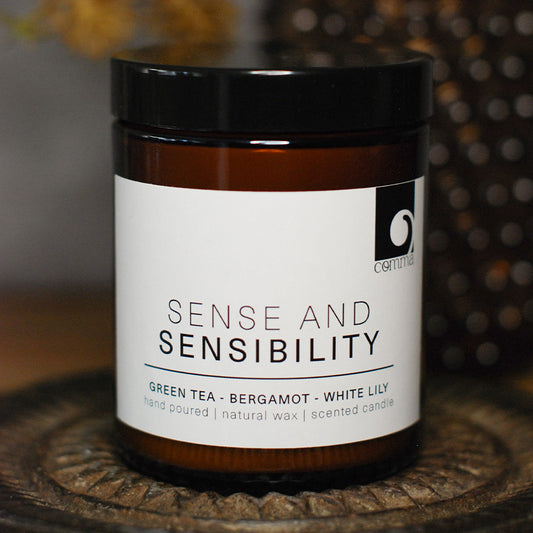 An amber glass jar candle with a black lid and a white label reading 'SENSE AND SENSIBILITY' and 'GREEN TEA - BERGAMOT - WHITE LILY'. The candle is photographed close-up on a textured round surface with a softly blurred background.